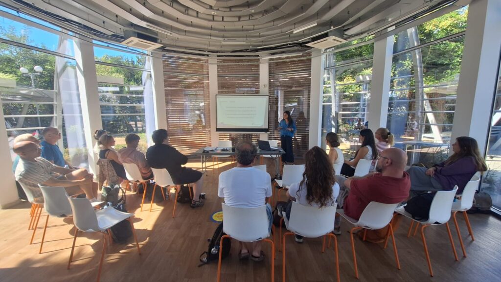 audience in a room , a screen in the middle, a woman standing next to it, explaining something